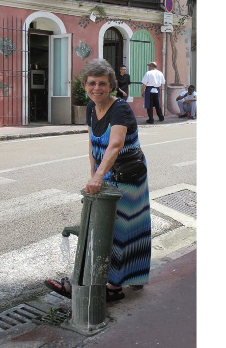 Photo shows Dona with her foot wearing a sandal under the water of a pump on the sidewalk of a narrow street.
